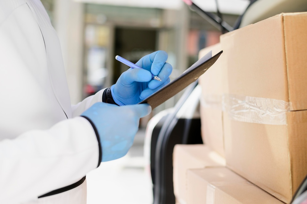 A person in a white coat and blue gloves holds a clipboard, likely in a medical or laboratory setting.